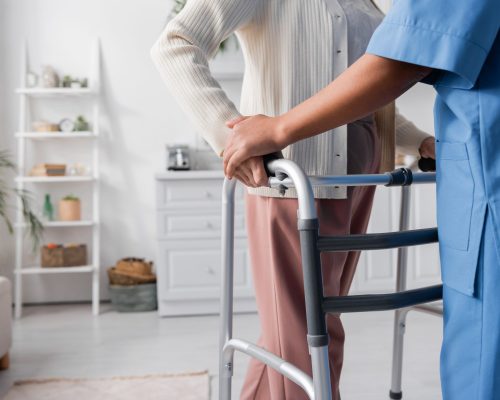 partial view of multiracial caregiver in uniform supporting senior woman walking with walker at home,stock image